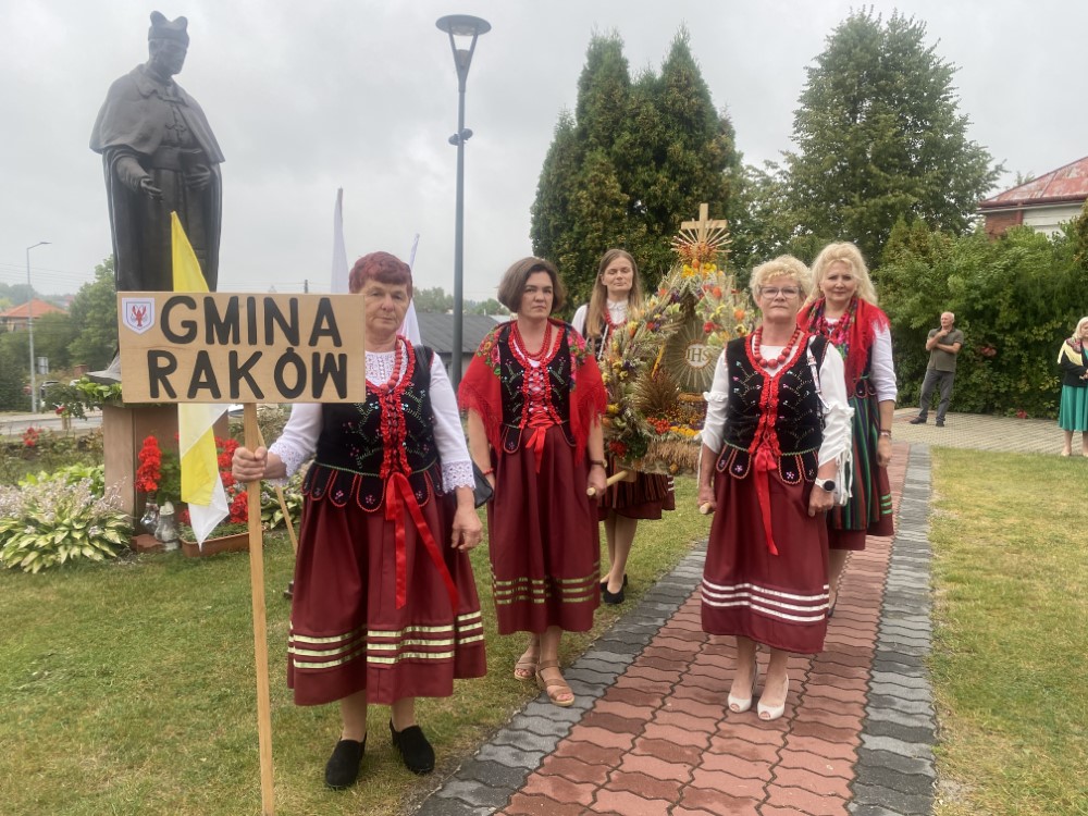 Kobiety ubrane w stroje ludowe, wieniec dożynkowy, tablica z napisem Gmina Raków