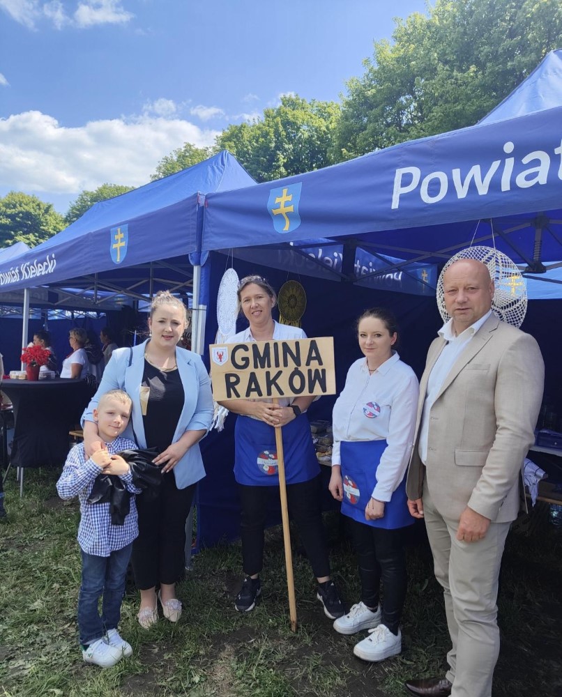 Osoby na tle namiotów handlowych, osoba pośrodku trzyma tablicę z napisem "GMINA RAKÓW"