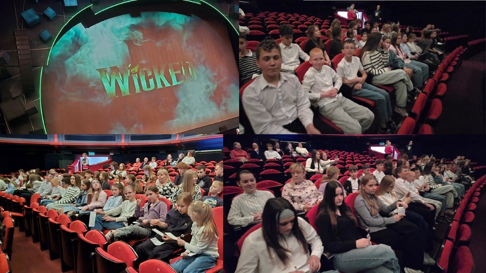 Widownia kina wypełniona dziećmi i młodzieżą siedzącymi w czerwonych fotelach przed seansem musicalu „Wicked”; na ekranie widoczny tytuł spektaklu w zielonej, dymnej oprawie.