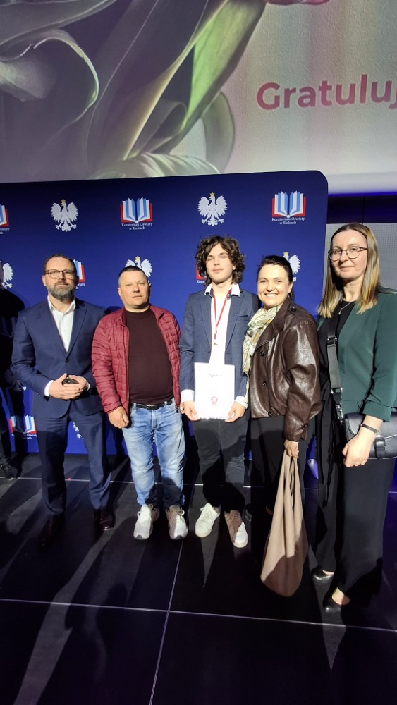 Pięć osób pozuje do zdjęcia na scenie podczas uroczystości wręczenia wyróżnienia. W centrum stoi młody mężczyzna trzymający dyplom i medal, a w tle widoczne są logotypy Kuratorium Oświaty.