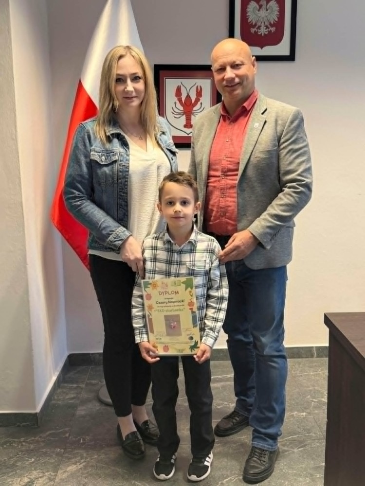 Kobieta, Wójt Gminy Raków i chłopiec pozują do zdjęcia w urzędzie. Chłopiec trzyma dyplom, a w tle widoczna jest flaga Polski oraz herby na ścianie.