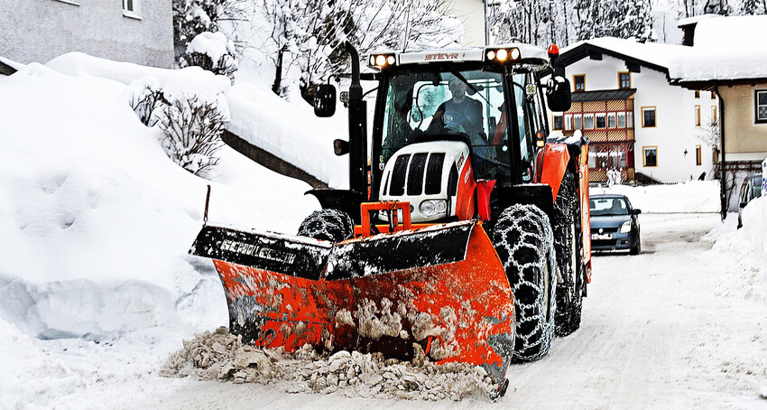 Na pierwszym planie traktor z pługiem do odśnieżania, w tle zaspy śniegowe i domy